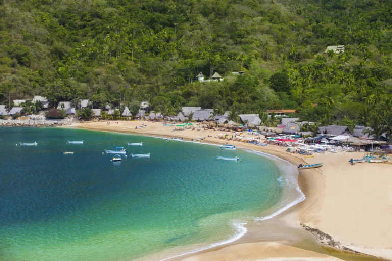 Small boats sit on calm water in front of a beach village built up a steep green hillside.